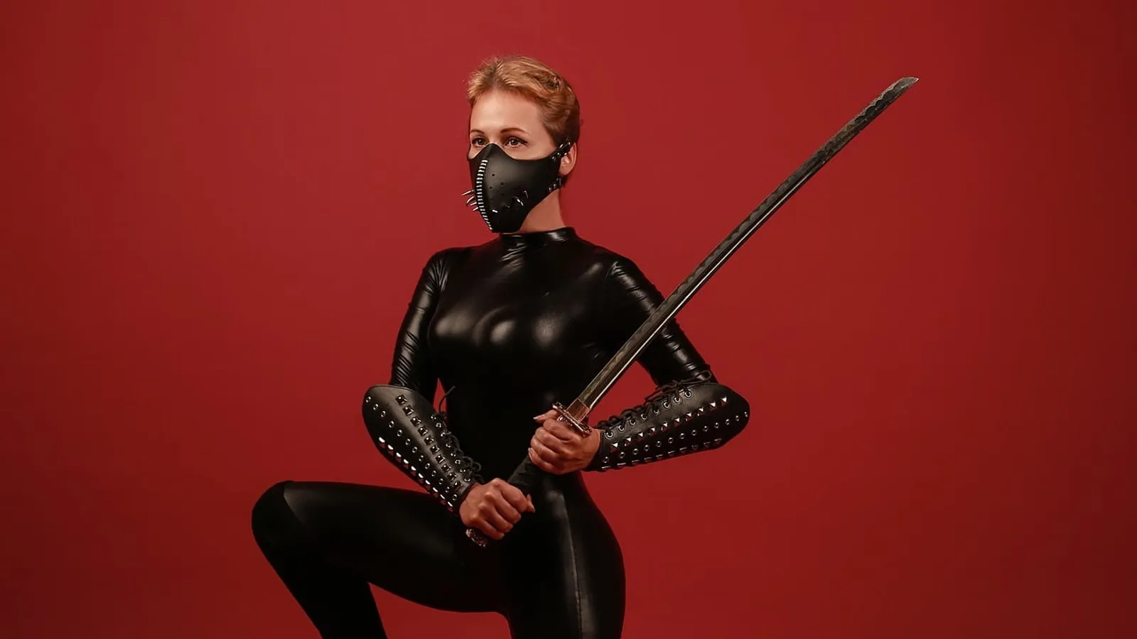 Kvinna i svart latexdräkt och mask mot röd bakgrund